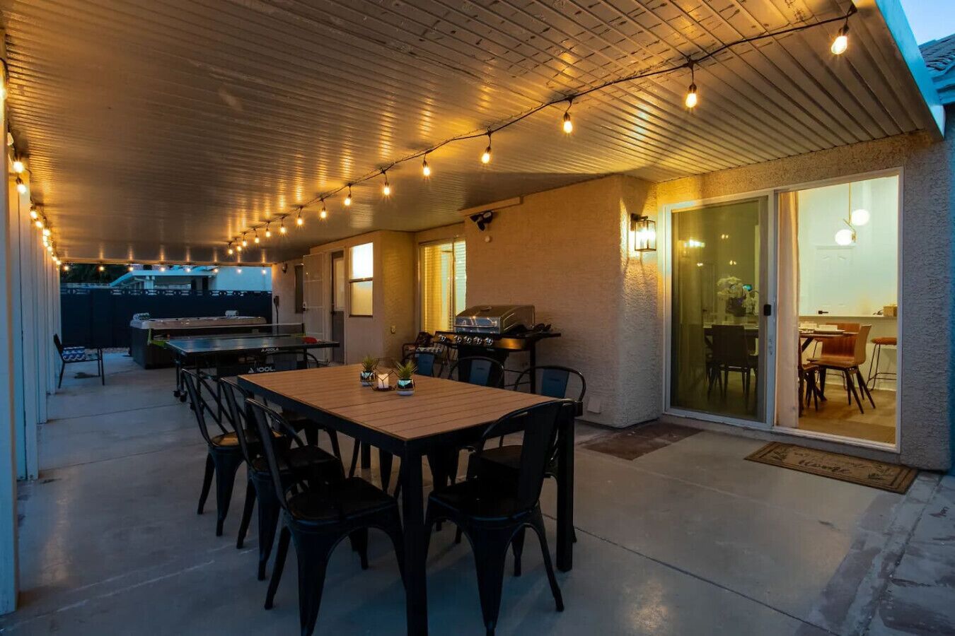 An outdoor patio with a wooden table and chairs, string lights, a grill, and a sliding glass door leading to an indoor area.