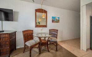 Un coin repas avec une table ronde en verre, deux chaises en osier, un meuble en bois, un téléviseur, un miroir et un tableau coloré accroché au mur.