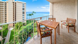 A balcony with a wooden table and chairs, overlooking a view of the ocean and palm trees. - Royal Kahana Maui 719 Ocean View Studio (Lahaina)