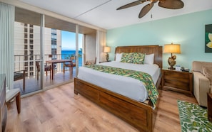A hotel room with a large bed, a balcony with a table and chairs, a ceiling fan, and a view of the ocean.