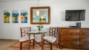 A dining area with a round glass table, two wooden chairs, a wooden cabinet, a television, and three landscape paintings on the wall. - Royal Kahana Maui 719 Ocean View Studio (Lahaina)