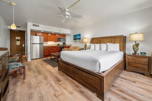 Une chambre d’hôtel avec un lit en bois, un bureau, une chaise, une table de chevet, une lampe, une horloge, un ventilateur de plafond et un coin cuisine équipé d’un réfrigérateur, d’un four à micro-ondes et d’une cuisinière.
