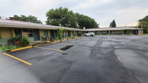 Een motel met een parkeerplaats, een witte auto geparkeerd op de plaats, en een rij motelkamers met groene luifels.