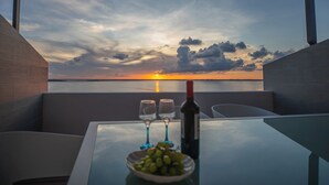 Et bord med en vinflaske, to glas og en skål med druer, med udsigt til solnedgang fra en balkon.