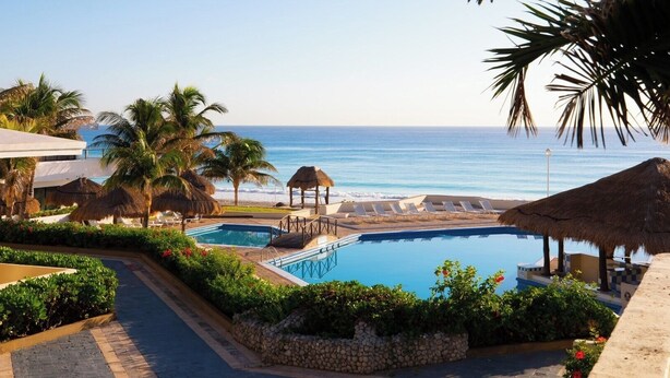 A resort pool area with a view of the ocean, palm trees, and a thatched-roof structure. - Exclusive Pent House, stunning ocean and lagoon views (Cancún)