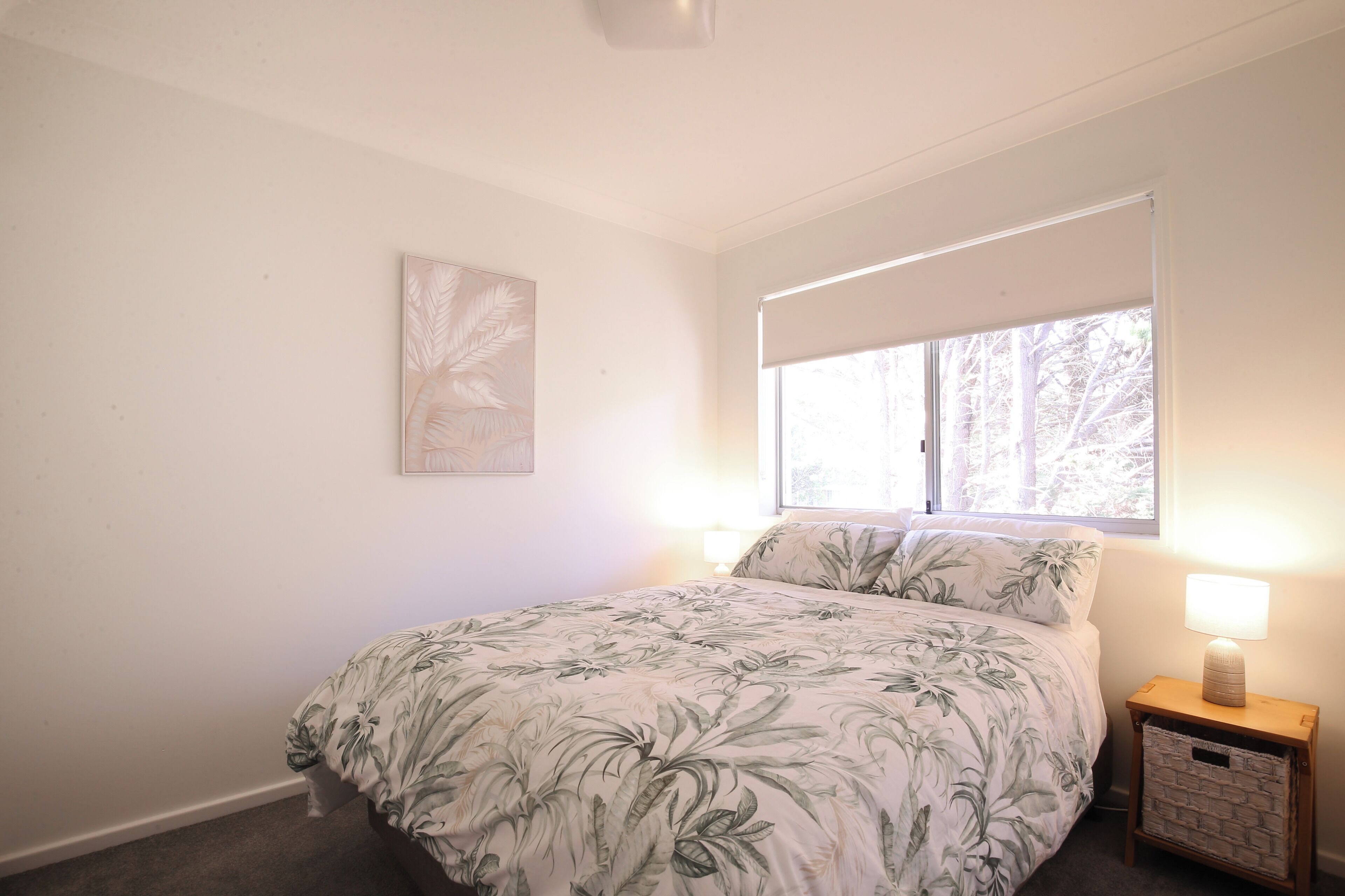A bedroom with a bed, a nightstand with a lamp, a window, and a wall-mounted artwork.