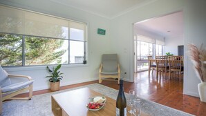 Ein modernes Wohnzimmer mit einem Holztisch, einer Obstschale, einer Weinflasche und Gläsern. Ein Sessel, ein Blumentopf und durch große Fenster ein Blick auf einen Essbereich sind ebenfalls zu sehen.