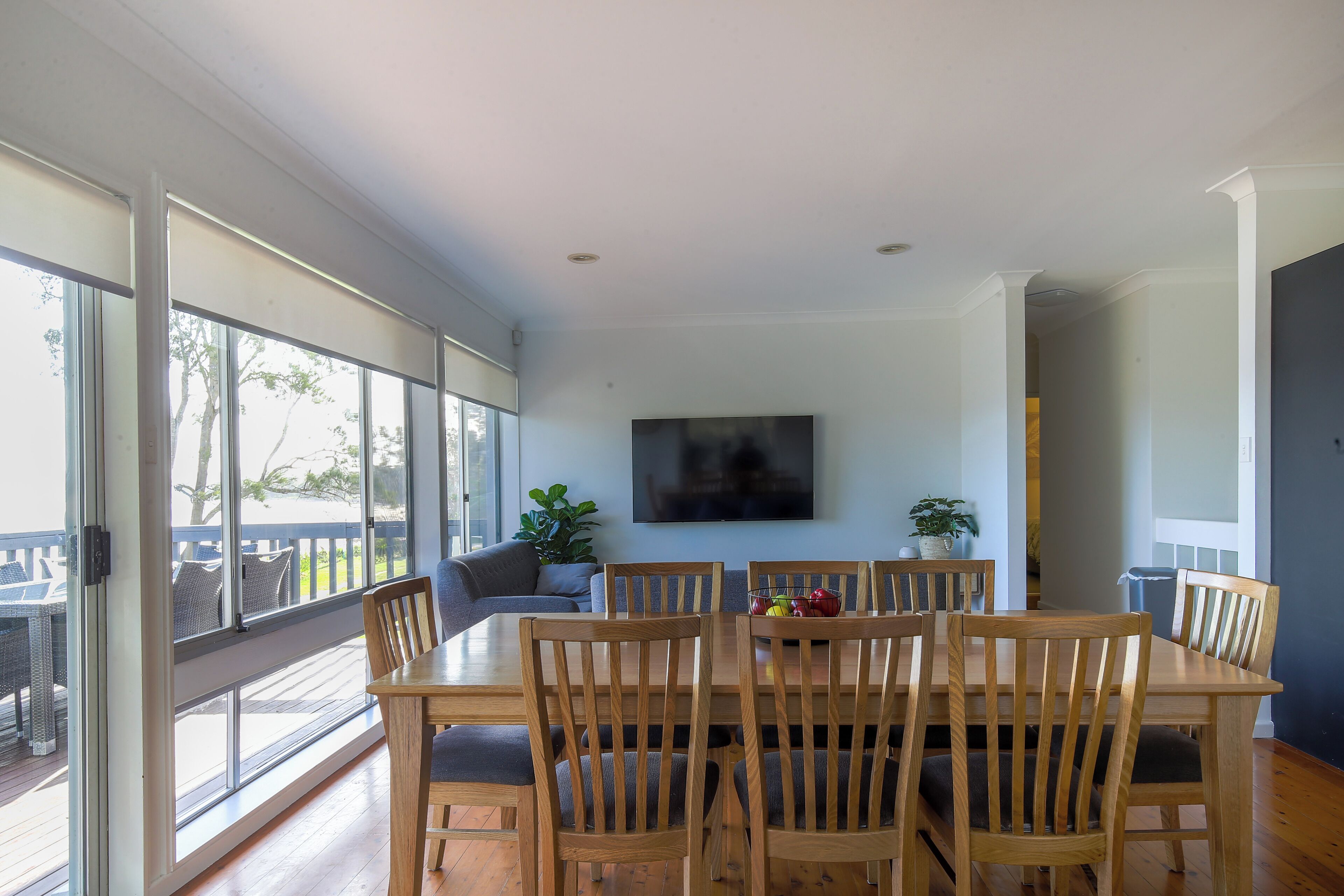 A dining area with a wooden table and chairs, a flat-screen TV mounted on the wall, and large windows offering a view of the outdoors.