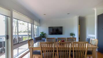 A dining area with a wooden table and chairs, a flat-screen TV mounted on the wall, and large windows offering a view of the outdoors.