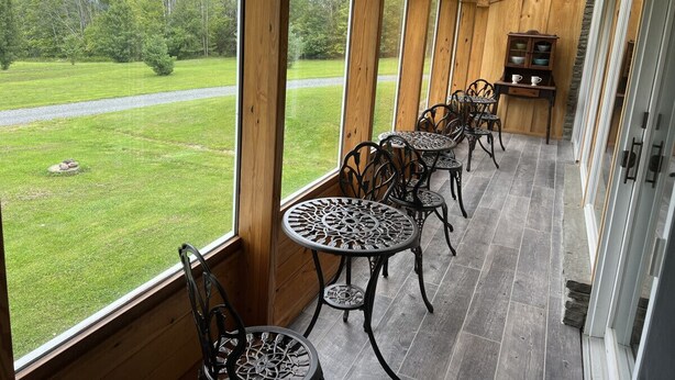 Un espace extérieur abrité, doté de murs en bois et de grandes fenêtres, aménagé avec des tables et des chaises en métal, ainsi qu’une cheminée en pierre.