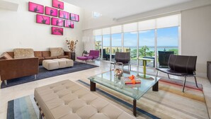A spacious living room with a large glass coffee table, a sofa, and multiple chairs. There are large windows offering a view of the outdoors.
