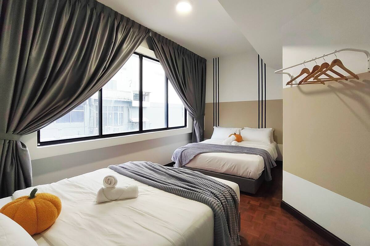 A hotel room with two beds, a large window with curtains, a wooden hanger rack, and a view of buildings outside.