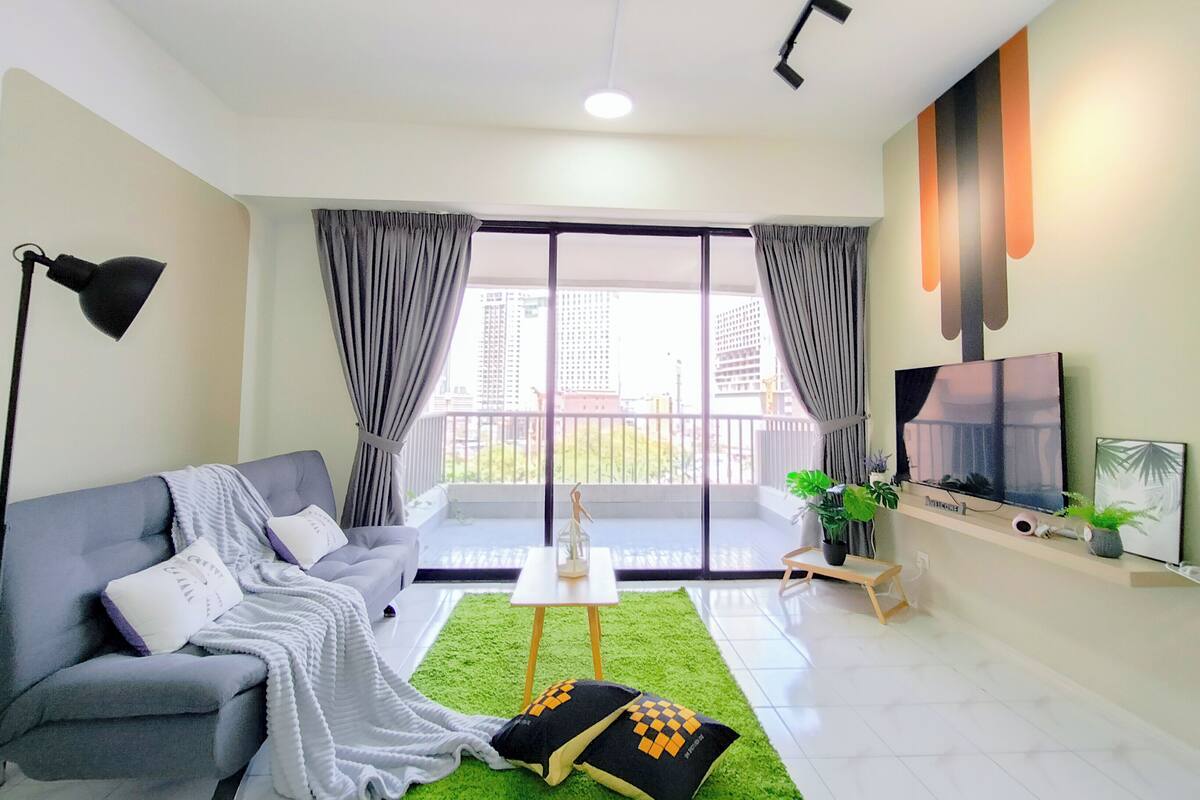 A modern living room with a grey sofa, a green rug, a flat-screen TV, and a balcony with curtains.