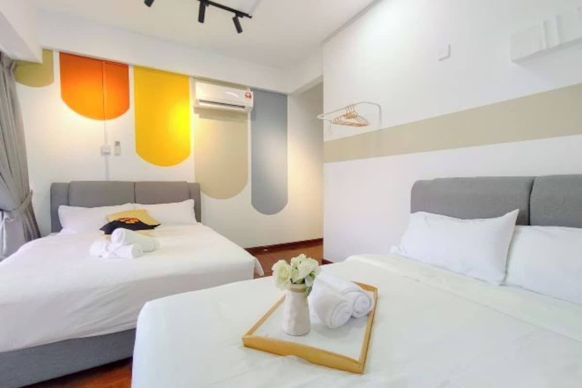 A hotel room with two beds, a wall-mounted air conditioner, and a bedside table with a tray of towels and a vase of flowers.