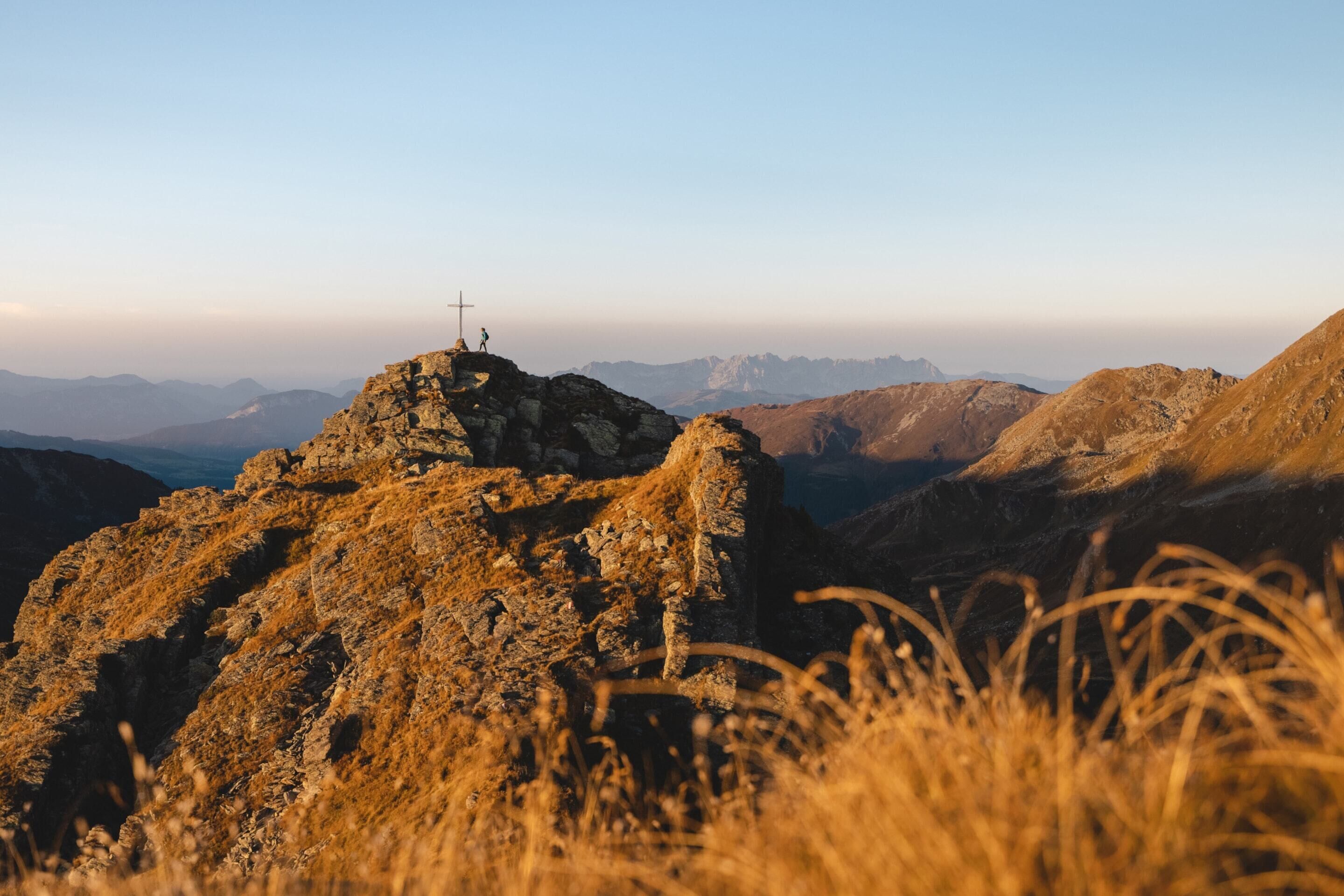 A rocky mountain peak with a cross at the summit and a vast mountainous landscape in the background.