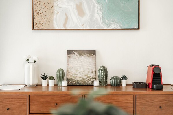 A wooden desk with a framed beach painting, a cactus, a small potted plant, a white vase with flowers, and a red object.