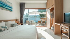 A modern hotel room with a large bed, a desk with a TV, and a balcony with outdoor seating.