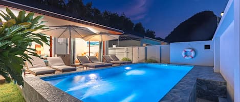 Une piscine entourée de chaises longues et de parasols, le tout bordé de murs en pierre et d’une pelouse.