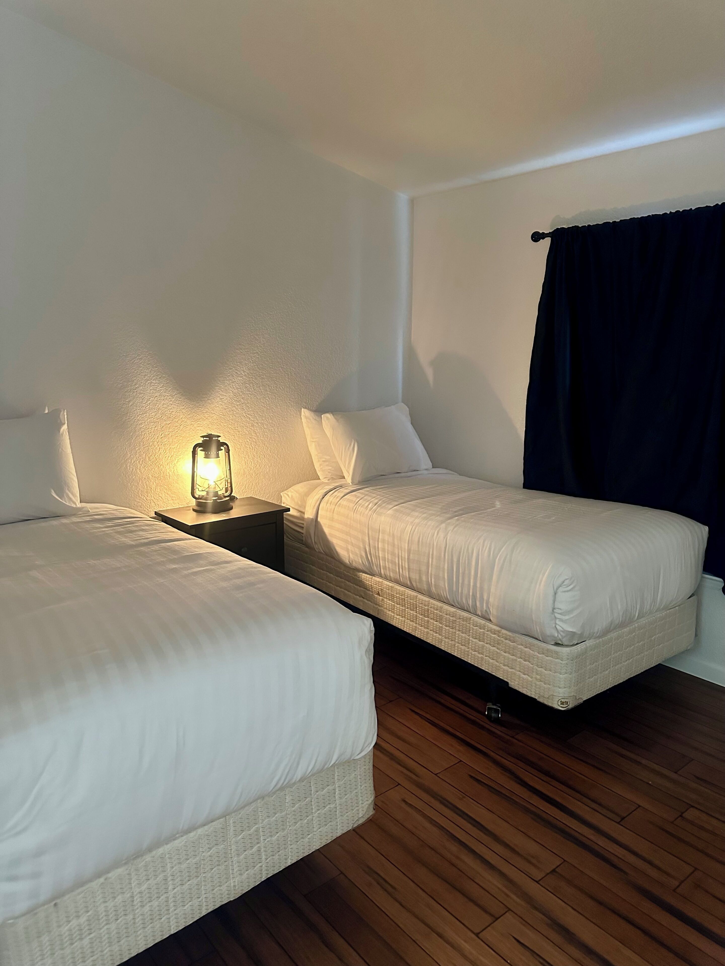 Ein Schlafzimmer mit zwei Betten, einem Nachttisch mit Lampe, Holzboden und einem dunklen Vorhang.