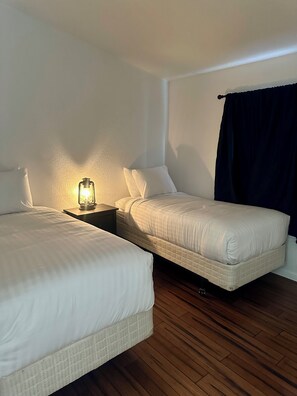 Ein Schlafzimmer mit zwei Betten, einem Nachttisch mit Lampe, Holzboden und einem dunklen Vorhang.