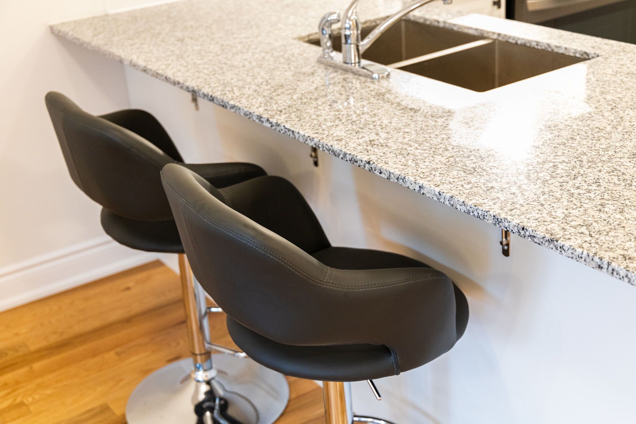 Eine Küche mit Granitarbeitsplatte und Spüle aus Edelstahl. Zwei schwarze Barhocker mit Metallbeinen sind vorhanden.