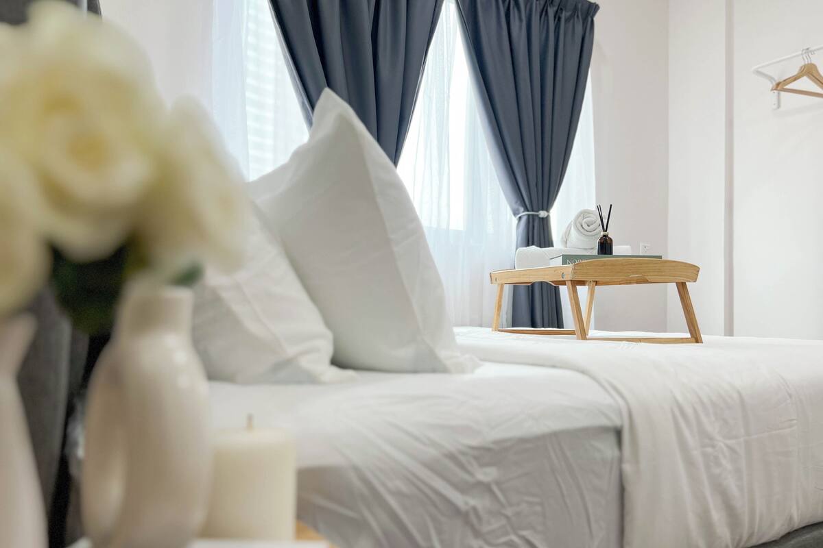 A neatly made bed with white linens, a bedside table with a book and a bottle, and a window with dark curtains.