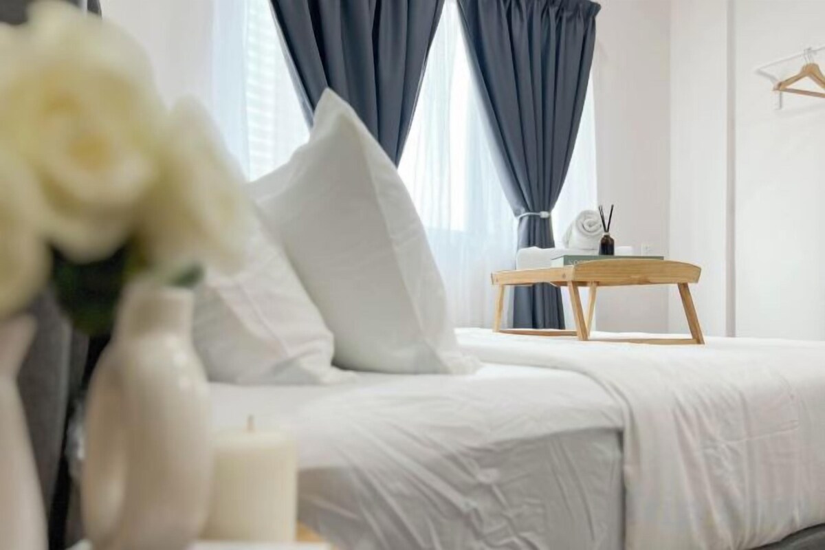 Uma cama com roupa de cama branca, uma mesa de cabeceira com abajur e um vaso com flores.