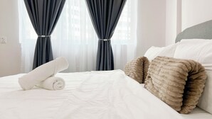A neatly made bed with white linens and rolled towels, dark curtains, and a grey headboard. - MH Maison by Manhattan Group  (Kuala Lumpur)
