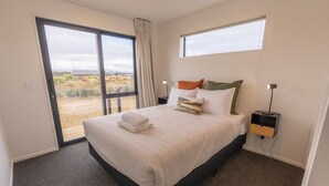 A bedroom with a bed, a nightstand, a lamp, and a view of the outdoors through a large window. - Tussock Bend Horizons (Twizel)