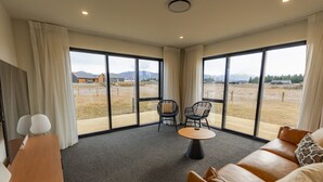 A spacious room with a large window, a leather sofa, two chairs, a small table, and a wooden desk. - Tussock Bend Horizons (Twizel)