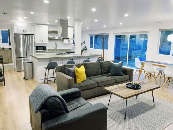 Relax in a spacious living area with a sofa, coffee table, and potted plant, adjacent to a kitchen with a dining area and large windows offering views of the outdoors.