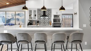 Une cuisine moderne avec un plan de travail blanc, un dosseret à motifs noir et blanc et un plafond en bois. On y trouve quatre tabourets de bar gris et deux suspensions.