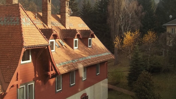 Una casa de dos pisos con fachada roja y techo de tejas, rodeada de un bosque y montañas.
