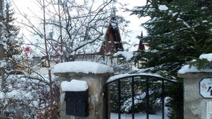Un portón de piedra con un portón metálico, rodeado de árboles cubiertos de nieve y un edificio con techo rojo al fondo.