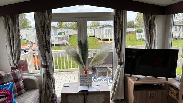 Un salon confortable avec un canapé, une table basse sur laquelle se trouvent un livre et un panneau, un téléviseur, et une vue sur une terrasse et des maisons mobiles à l’extérieur.