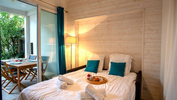 A hotel room with a bed, a dining area with a table and chairs, a lamp, and a view of greenery outside.