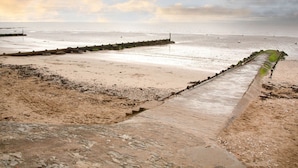 A coastal beach with a wooden walkway leading to the sea, a rocky foreground, and a cloudy sky.