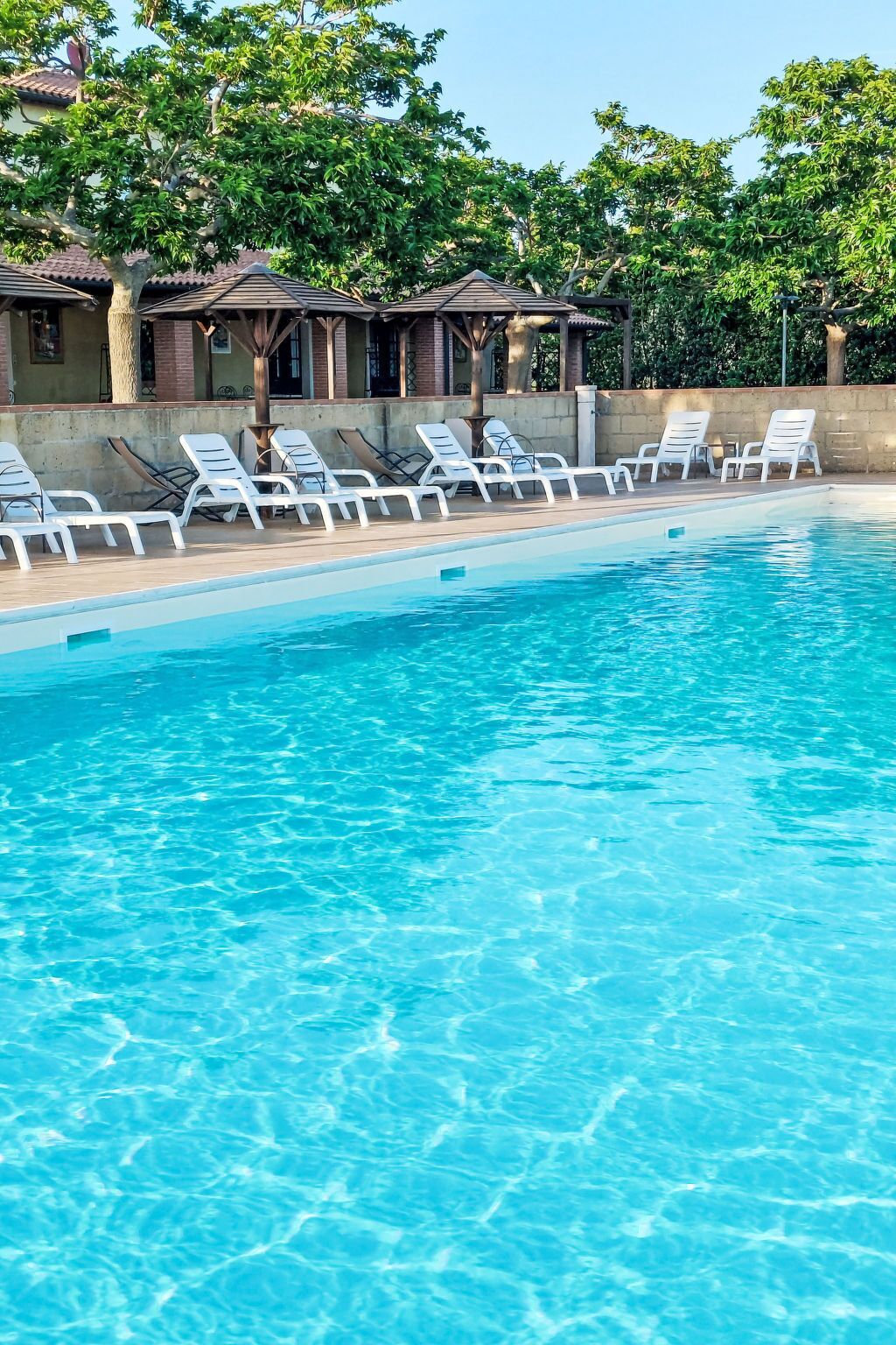 Une piscine entourée de chaises longues blanches et d’un espace abrité avec des tables et des parasols.