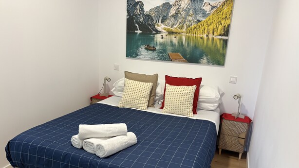Een netjes opgemaakt bed met een blauw geruit beddegoed en twee opgerolde witte handdoeken, een schilderij van een meer en bergen aan de muur, en een houten vloer.