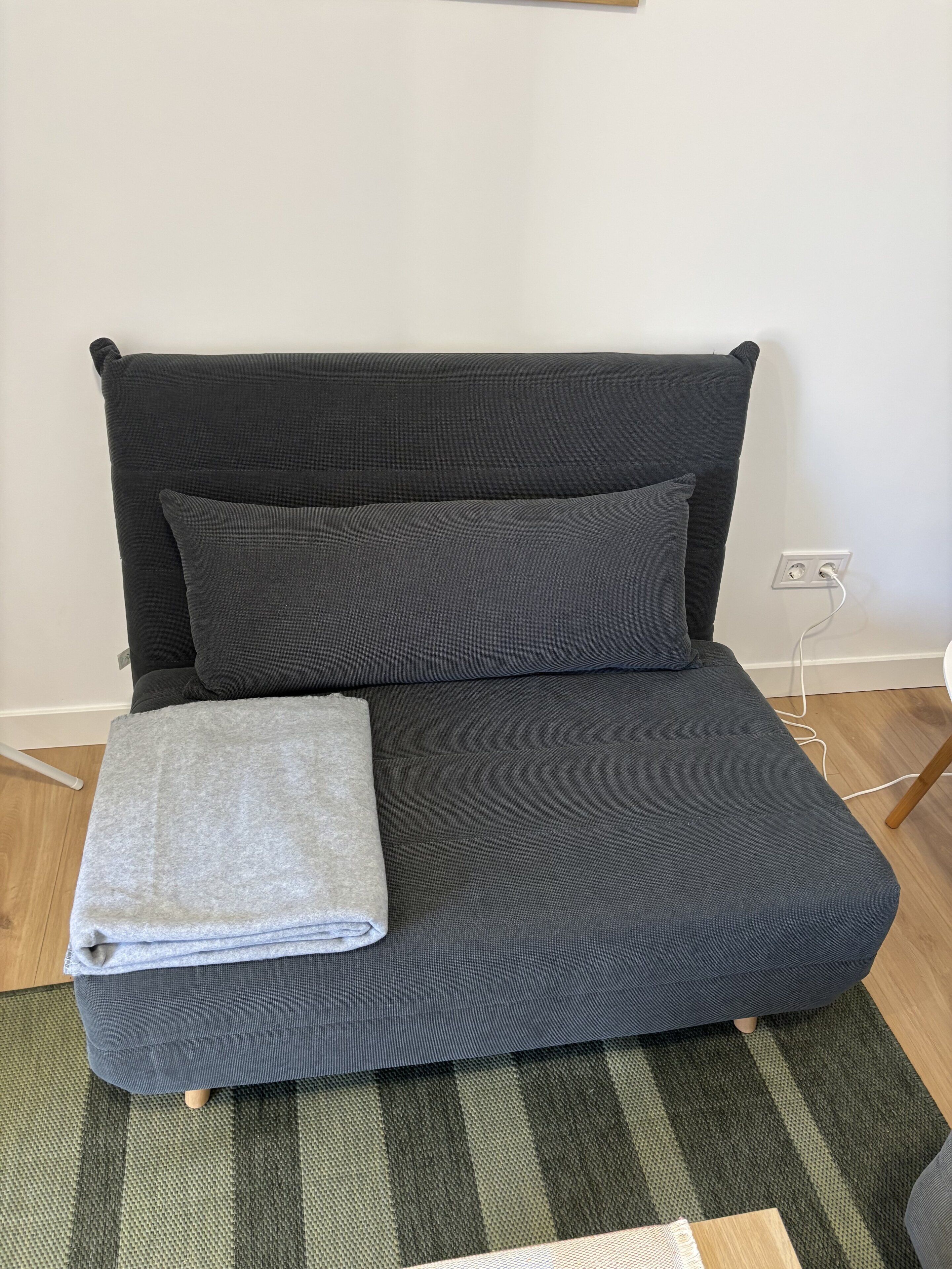 Ein Bett mit dunklem Kopfteil, Kissen und einer zusammengefalteten Decke. Eine Steckdose ist an der Wand zu sehen.