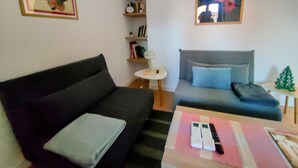 Een woonkamer met een zwarte bank, een grijze zithoek, een klein rond tafeltje en een boekenkast.