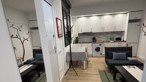 Una cocina moderna con armarios blancos, un microondas y una cafetera. Hay una zona de comedor con una mesa y sillas, y un sofá al fondo.