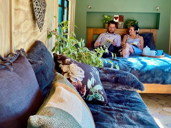 Une pièce chaleureuse avec un canapé et un lit, tous deux dotés de linge de lit bleu. On trouve des plantes en pot sur le lit et une étagère murale agrémentée d’objets décoratifs.