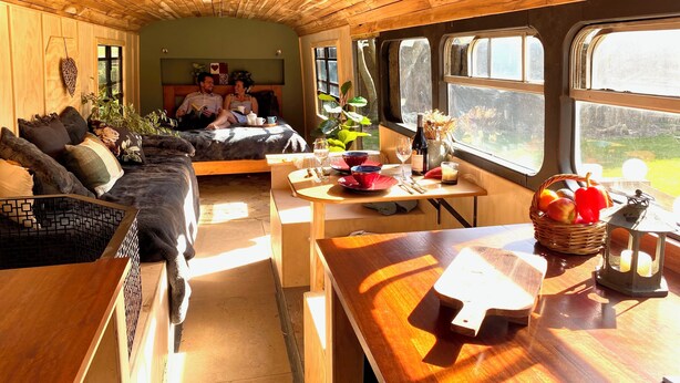 Ein gemütliches Blockhäuschen mit Holzbalkendecke, einer Couch, einem Esstisch mit Obstkorb und einem Fenster mit Blick auf einen Garten.