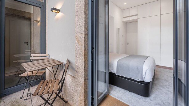 A modern bedroom with a bed, a small table, and a bench. There is a sliding door leading to another room. - GuestReady - Mompilher Apartments - D (Porto)