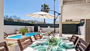 An outdoor dining area with a pool, wooden chairs, and a table set for a meal. - V2 Caliços - VIP Properties by Interhome (Albufeira)