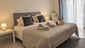 A neatly made bed with a grey headboard, white and black pillows, and rolled towels. There is a small table with a lamp beside the bed, and a chair with a hat on it. - V2 Caliços - VIP Properties by Interhome (Albufeira)