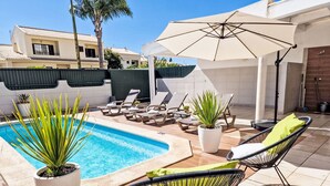 A swimming pool with lounge chairs and umbrellas, surrounded by potted plants and a tiled patio area. - V2 Caliços - VIP Properties by Interhome (Albufeira)