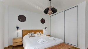 A modern bedroom with a wooden bed, a wooden bench, two bedside tables, a large window, and two wall-mounted light fixtures. - Chanquete 2310  One Bedroom (Manilva)