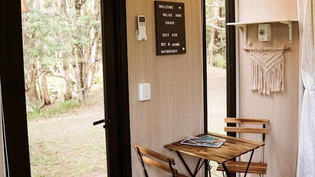 Una habitación con una mesa y sillas de madera, una puerta que da al exterior, un letrero de bienvenida y un tapiz de macramé en la pared.